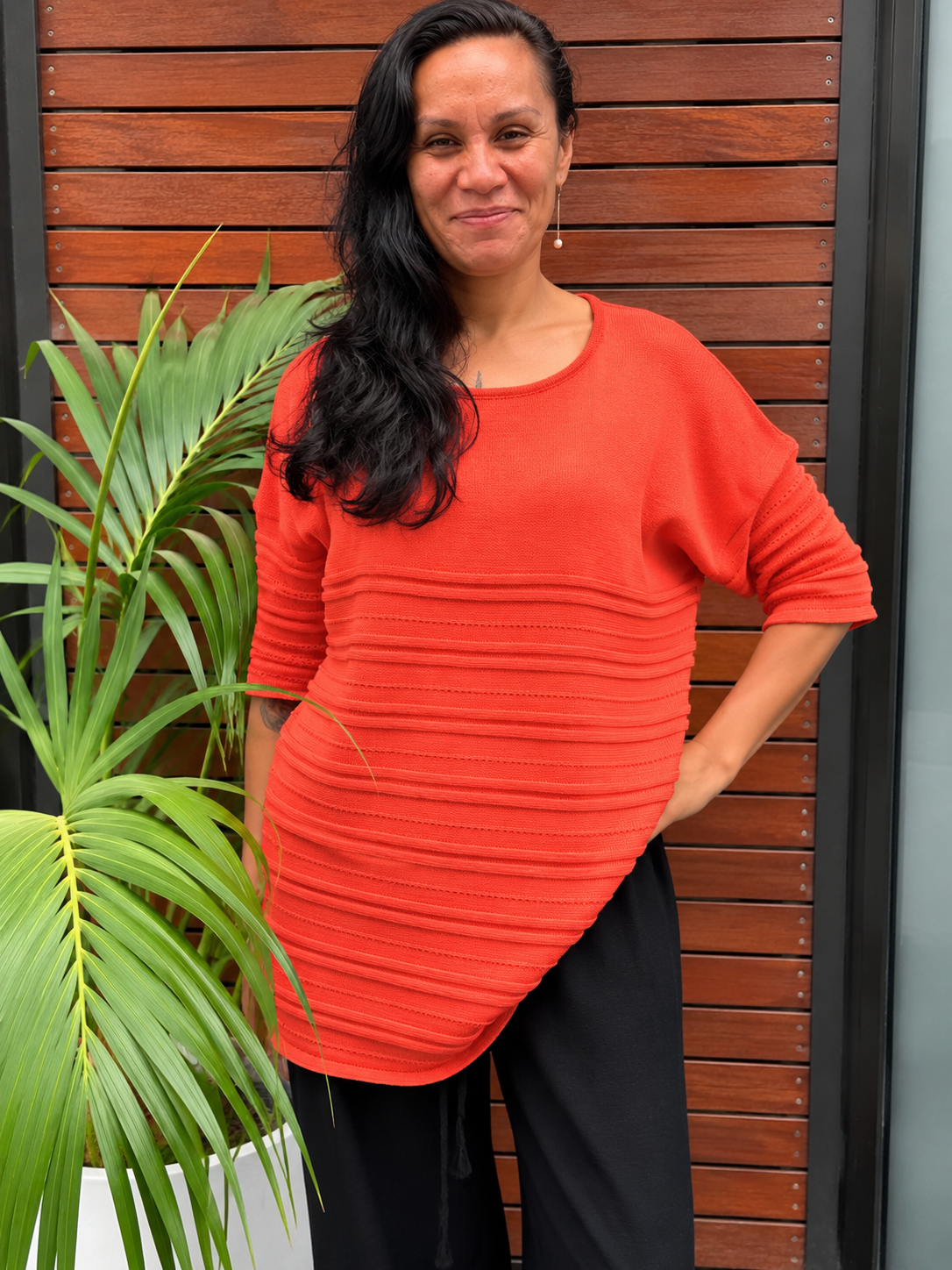 A woman wearing a bright orange ribbed knit top with elbow-length sleeves and black pants, standing in front of a brown wooden slat wall next to a green potted plant.