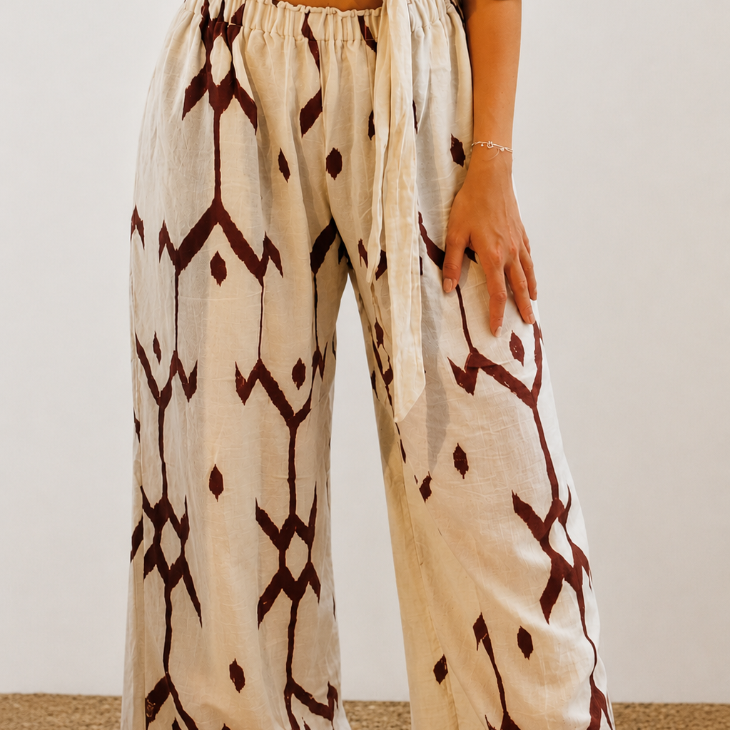 A person wearing loose, wide-leg pants with an off-white base and dark brown geometric ikat-style patterns, paired with a cream tie-front top. The model is standing barefoot on a textured tan rug against a plain off-white wall.