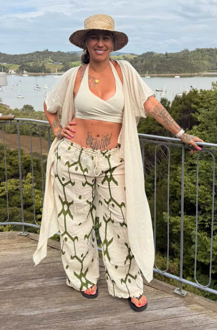 A woman stands on a wooden deck outdoors by the water wearing a straw hat, a cream crop top, a matching open-front cardigan, and cream wide-leg pants with a green botanical print.